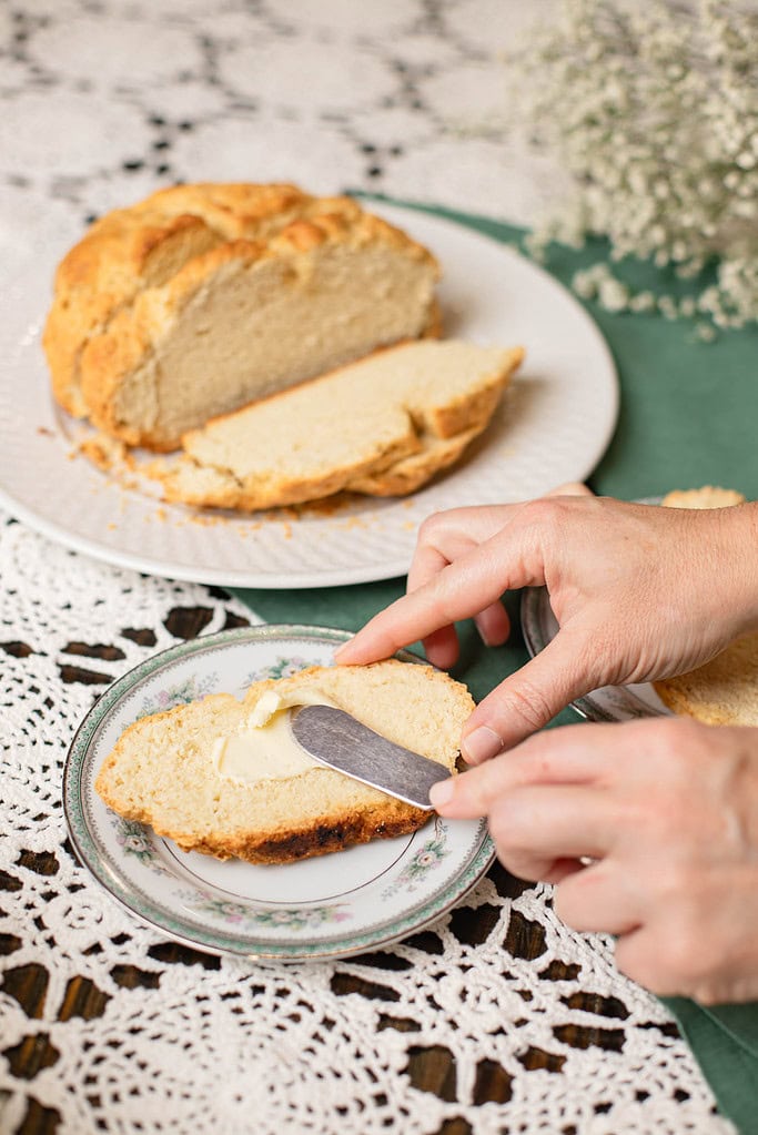 Hands spreading butter onto a slice of warm Irish soda bread on a small plate, with the sliced loaf visible in the background.