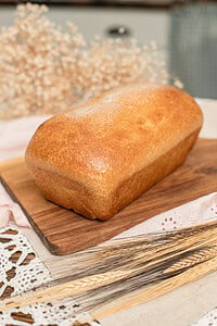 Whole unsliced fresh milled sourdough sandwich bread loaf with a deep golden-brown crust on a wood cutting board, styled with dried wheat stems on a pink eyelet linen and crochet lace, with a soft-focus cottage kitchen background.