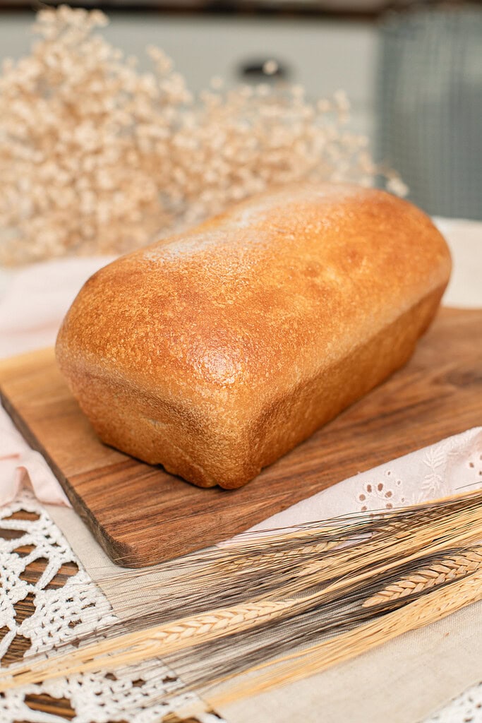 Whole unsliced fresh milled sourdough sandwich bread loaf with a deep golden-brown crust on a wood cutting board, styled with dried wheat stems on a pink eyelet linen and crochet lace, with a soft-focus cottage kitchen background.