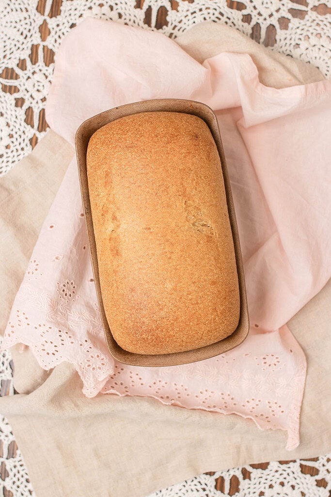 Overhead view of a freshly baked fresh milled sourdough sandwich bread loaf in a stoneware pan, showing a golden-brown rounded crust, nestled in a pink eyelet linen with crochet lace on a wood surface.