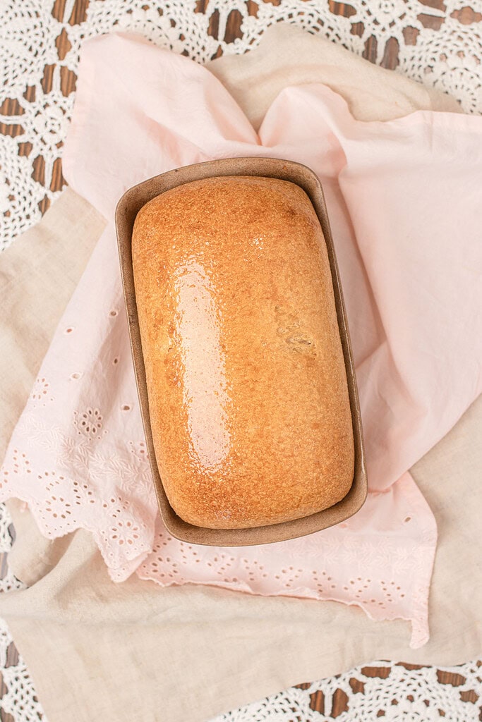 Overhead view of a fresh milled sourdough sandwich bread loaf in a stoneware pan, just out of the oven with a shiny butter-brushed golden crust, resting on a pink eyelet linen and crochet lace.
