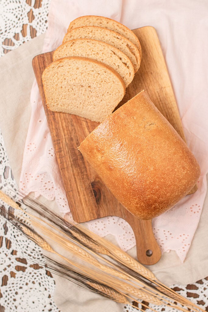 Overhead view of a partially sliced fresh milled sourdough sandwich bread loaf on a wood cutting board, with four slices fanned beside the remaining loaf, styled on a pink eyelet linen with dried wheat stalks and crochet lace.