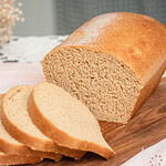 Close-up of a sliced fresh milled sourdough sandwich bread loaf on a wood cutting board, showing a fine, even golden crumb on the cut face with three slices leaning forward, styled on a pink eyelet linen with dried wheat stems and soft-focus dried botanicals in the background.