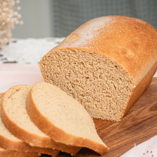 Close-up of a sliced fresh milled sourdough sandwich bread loaf on a wood cutting board, showing a fine, even golden crumb on the cut face with three slices leaning forward, styled on a pink eyelet linen with dried wheat stems and soft-focus dried botanicals in the background.