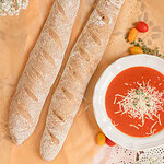 Two freshly baked sourdough baguettes made with freshly milled flour rest on parchment paper beside a white bowl of tomato soup topped with shredded cheese and herbs, styled with small tomatoes, greenery, and delicate white flowers on a rustic lace tablecloth.