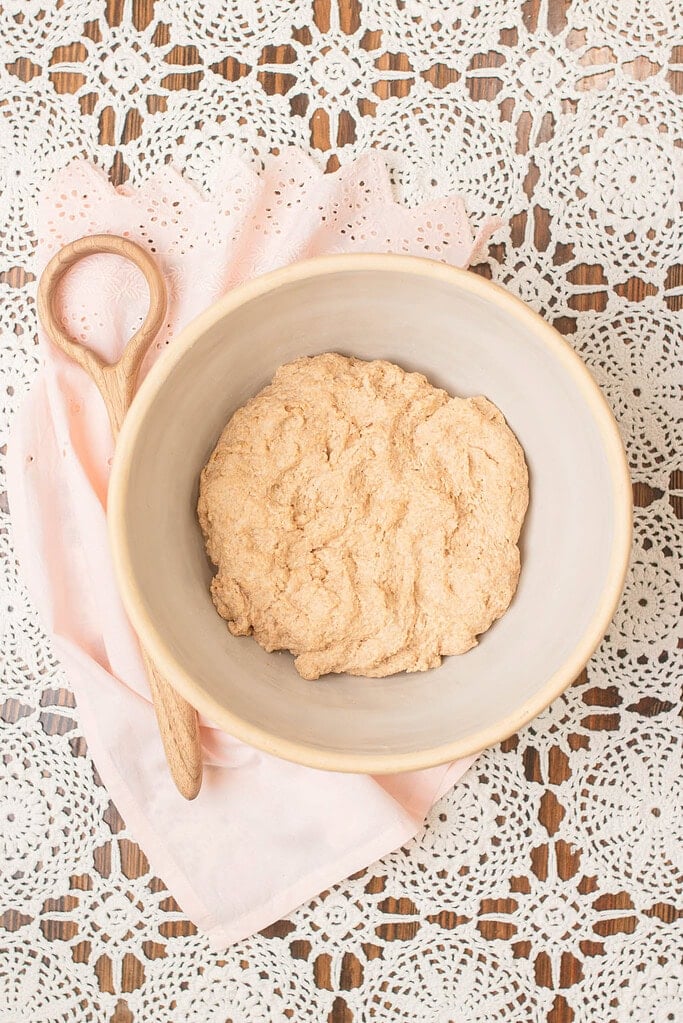 Freshly mixed sourdough dough made with freshly milled flour resting in a large ceramic bowl, styled with a wooden spoon and soft pink cloth on a detailed white lace tablecloth.