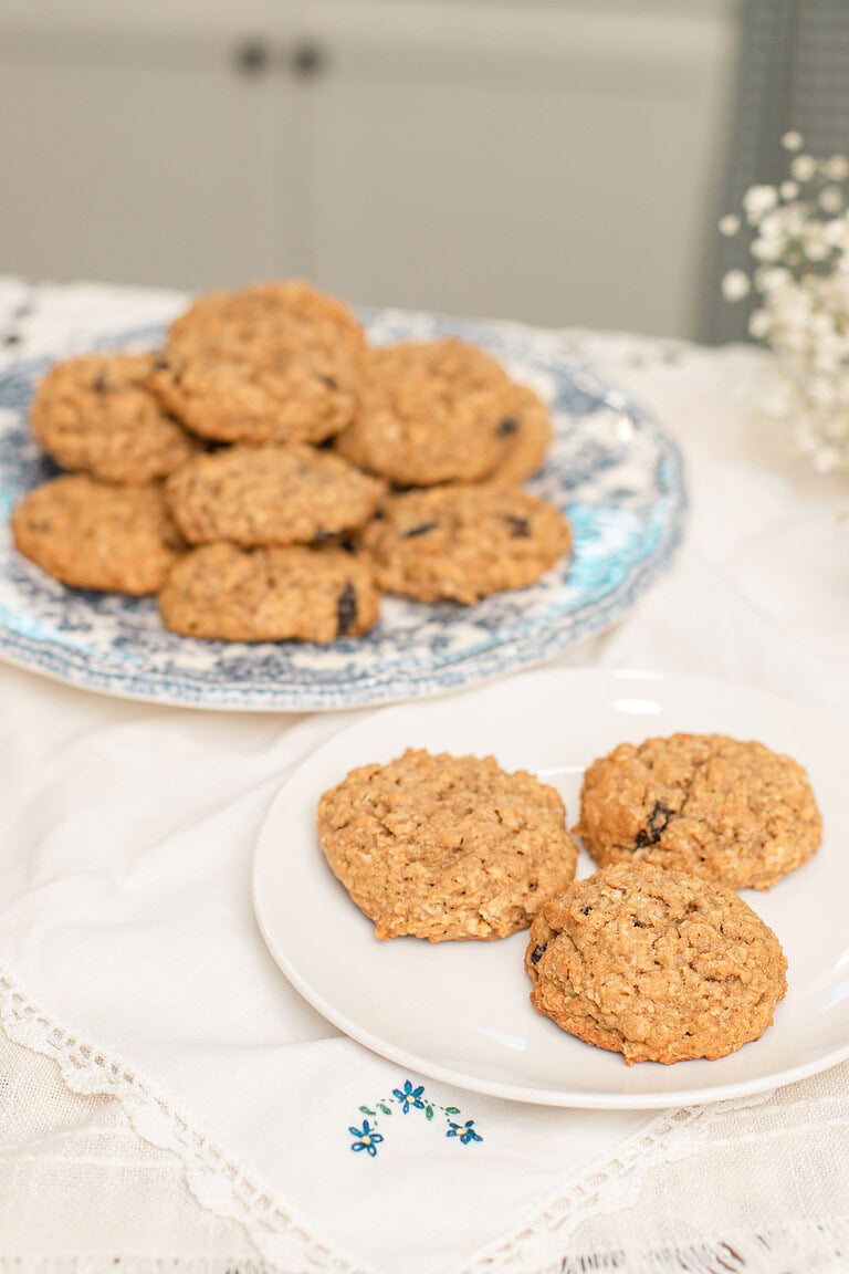 Sourdough einkorn oatmeal raisin cookies on a white plate in the foreground with three cookies, and a blue and white floral china plate stacked with more cookies in the background, styled on a white embroidered linen tablecloth with baby's breath flowers.
