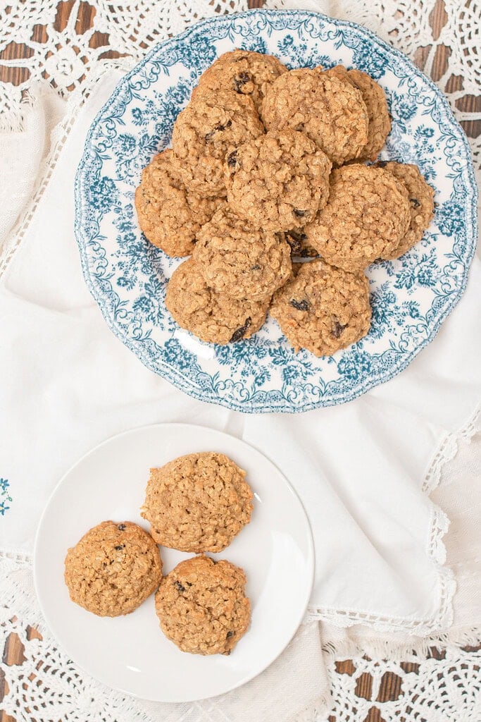 Overhead view of sourdough einkorn oatmeal raisin cookies piled on a blue floral china plate beside a small white plate with three cookies, styled on a white linen napkin and crochet lace on a wood surface.