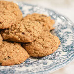 Close-up of sourdough einkorn oatmeal raisin cookies piled on a blue and white floral china plate, showing the golden oat texture and raisins, with soft white baby's breath flowers blurred in the background.