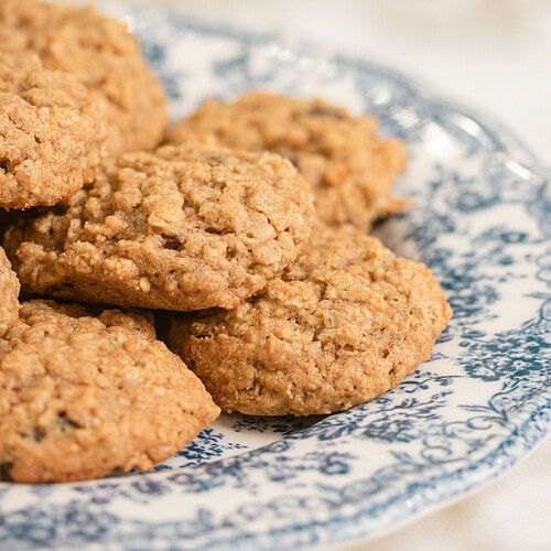 Close-up of sourdough einkorn oatmeal raisin cookies piled on a blue and white floral china plate, showing the golden oat texture and raisins, with soft white baby's breath flowers blurred in the background.