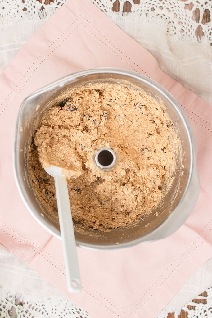Overhead view of finished sourdough einkorn oatmeal raisin cookie dough in a stand mixer bowl with a white spatula, showing oats and raisins folded throughout, on a pink linen napkin.