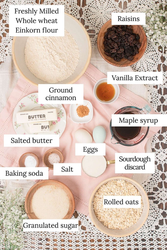 Labeled overhead ingredient flat lay for sourdough einkorn oatmeal raisin cookies showing freshly milled whole wheat einkorn flour, raisins, salted butter sticks, eggs, maple syrup, sourdough discard, rolled oats, granulated sugar, ground cinnamon, vanilla extract, baking soda, and salt on a pink eyelet linen with crochet lace and white baby's breath.