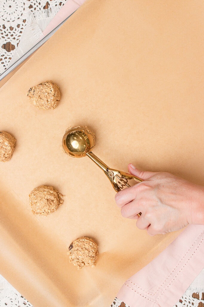 Hand using a gold cookie scoop to portion sourdough einkorn oatmeal raisin cookie dough onto a parchment-lined baking sheet, with several dough mounds already placed on the sheet.