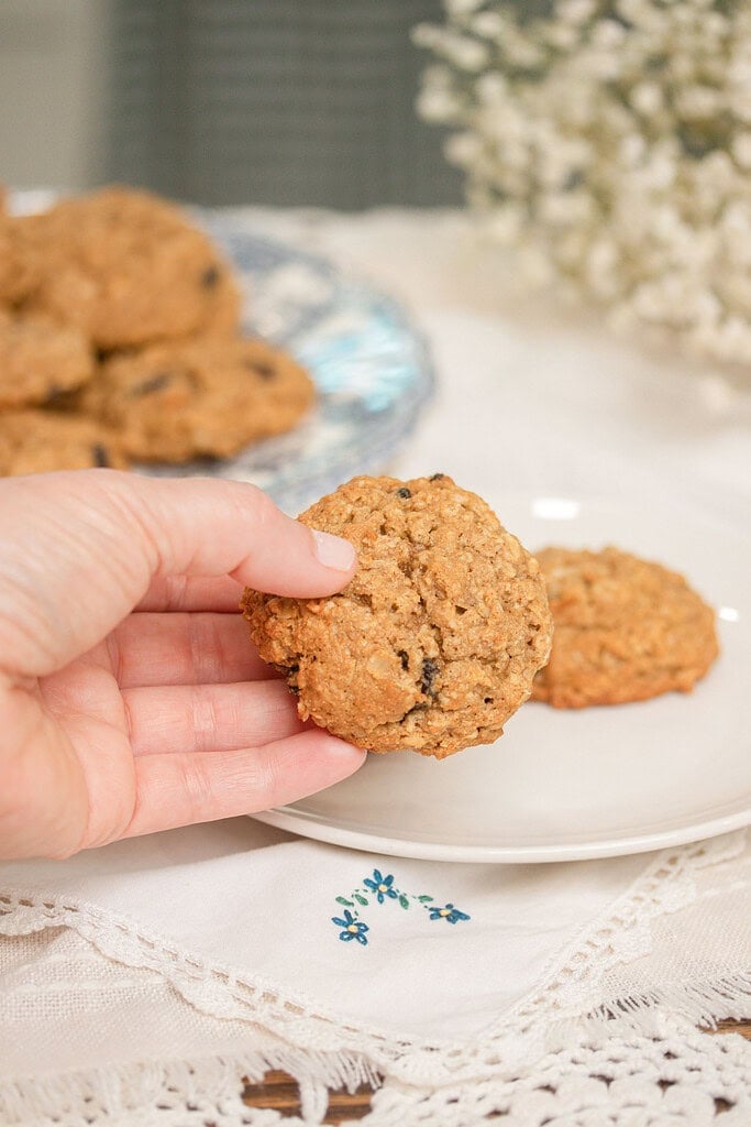 Hand holding a single sourdough einkorn oatmeal raisin cookie up close, showing the golden oat texture and raisins, with a white embroidered plate and more cookies blurred in the background alongside baby's breath flowers.