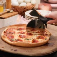 A woman is slicing a sourdough pizza on a ceramic pizza stone in a cottage kitchen on a wooden countertop.