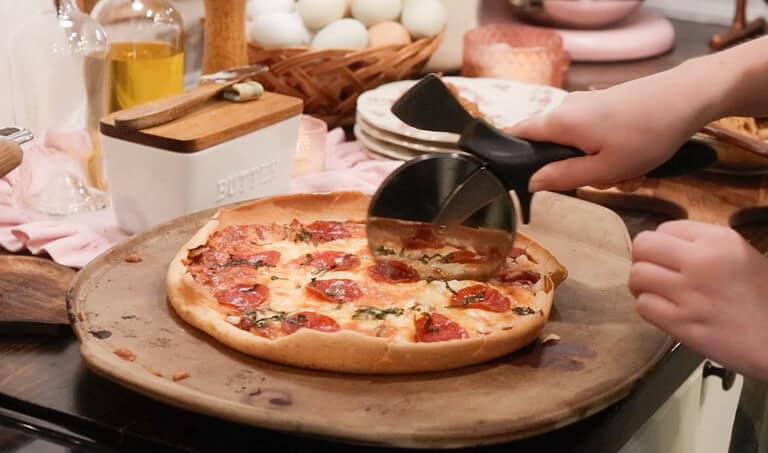A woman is slicing a sourdough pizza on a ceramic pizza stone in a cottage kitchen on a wooden countertop.