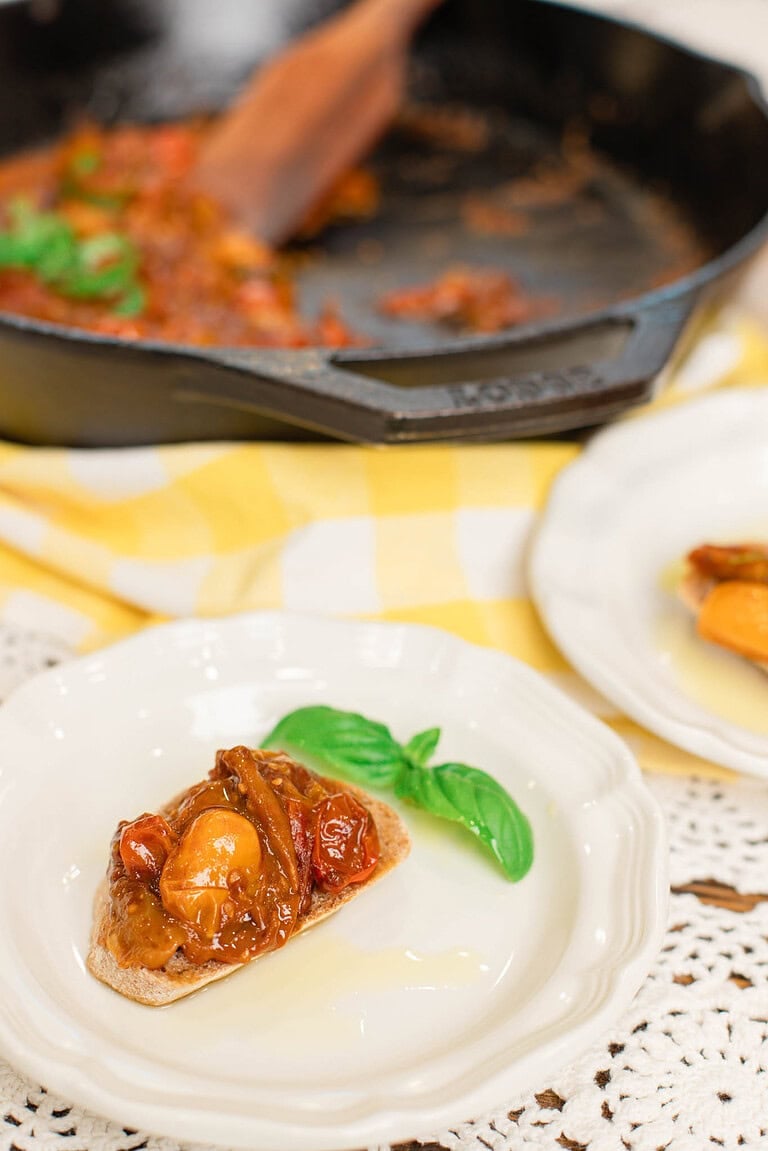 Slice of sourdough baguette topped with a rich tomato bruschetta mixture resting on a white plate with a fresh basil leaf, with a cast iron skillet of cooked tomatoes in the background on a lace tablecloth.