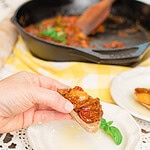 Hand holding a slice of sourdough baguette topped with glossy tomato bruschetta and garlic over a white plate with a basil leaf, with a cast iron skillet in the background.