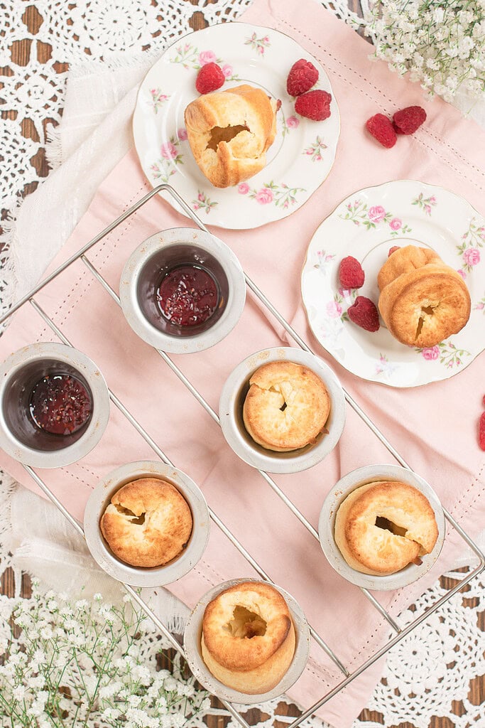 Overhead flat lay of raspberry sourdough popovers in vintage aluminum popover cups on a wire rack, with two popovers plated on rose-patterned china beside fresh raspberries, small bowls of raspberry jam, and white baby's breath flowers on a pink linen.
