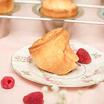 Single sourdough popover with a tall, golden-brown hollow crown on a rose-patterned vintage china plate, with fresh raspberries alongside and more popovers on the wire rack blurred in the background, on a pink linen surface.