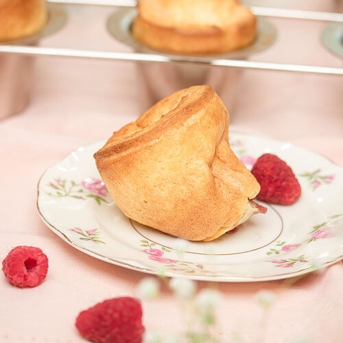 Single sourdough popover with a tall, golden-brown hollow crown on a rose-patterned vintage china plate, with fresh raspberries alongside and more popovers on the wire rack blurred in the background, on a pink linen surface.