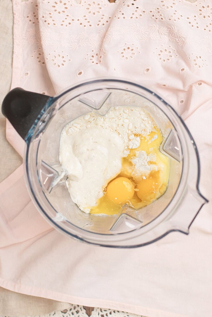 Overhead view of sourdough popover batter ingredients in a blender — sourdough starter, eggs, and milk — ready to blend, on a pink eyelet fabric background.