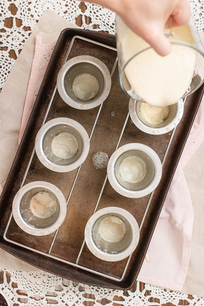 Hand pouring sourdough popover batter from a glass measuring cup into preheated aluminum popover cups on a dark baking sheet, with the remaining cups ready to fill, on a pink linen and crochet lace surface.