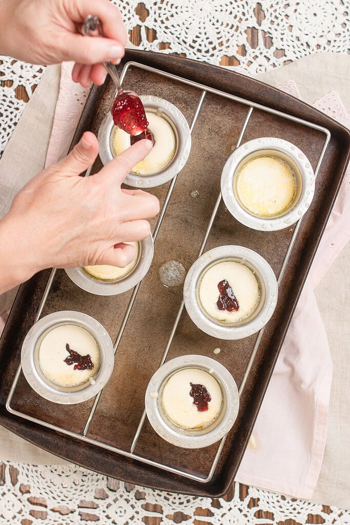 Hand spooning raspberry jam into sourdough popover batter in aluminum popover cups on a dark baking sheet, with jam already added to several cups and one spoonful being swirled in.