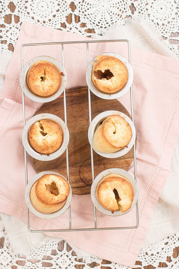 Overhead view of six freshly baked raspberry sourdough popovers in vintage aluminum popover cups on a wire rack, showing golden-brown tops with hollow centers, on a pink linen and crochet lace surface.
