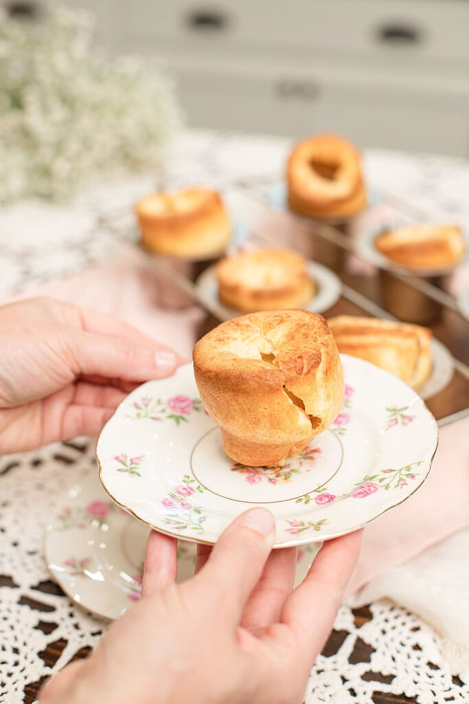 Two hands holding a vintage rose-patterned china plate presenting a single tall, golden sourdough popover with a hollow open crown, with the full popover rack blurred in the background on a pink linen surface.
