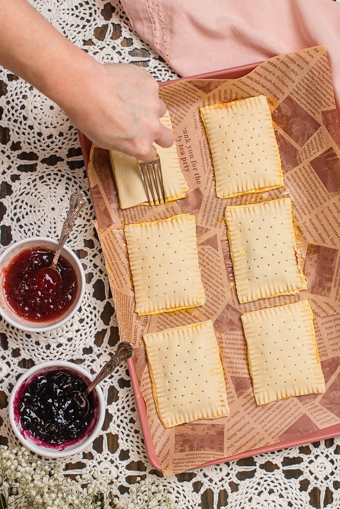 Crimping the edges of sourdough discard pop-tarts with a fork on a lined baking tray with jam nearby.