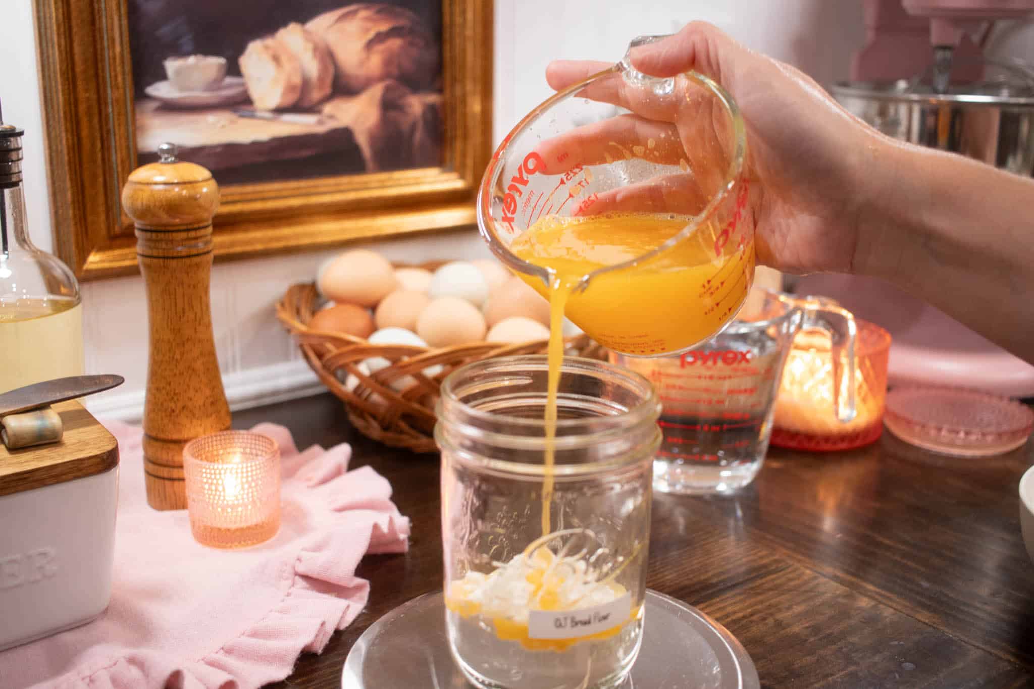 a woman pouring orange juice into a pint-sized glass mason jar on a kitchen scale on a wooden scale with two candles glowing in the background