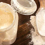 A clear glass flip top lid jar is holding sourdough starter in a cottage kitchen with a wooden scoop of bread flour beside it.