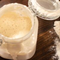 A clear glass flip top lid jar is holding sourdough starter in a cottage kitchen with a wooden scoop of bread flour beside it.