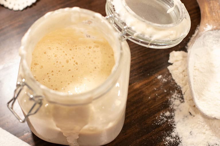 A clear glass flip top lid jar is holding sourdough starter in a cottage kitchen with a wooden scoop of bread flour beside it.