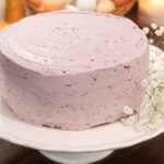 A phot of a finished blueberry cake with a fresh sprig of babys breath on a white pedestal cake stand in a cottage kitchen on a wooden counter top.
