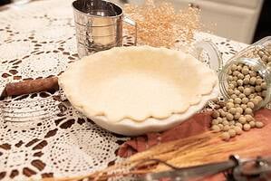 An unbaked sourdough pie crust in a ceramic pie dish, set on a lace tablecloth. A jar of pie weights spills out beside the dish, alongside vintage kitchen tools and dried floral decor, creating a cozy, rustic baking scene.