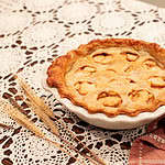 Another angle of the baked sourdough apple pie with a golden, flaky crust, showcasing the apple slices visible through the cutouts, styled with wheat stalks and vintage baking tools on a lace doily.