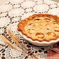Another angle of the baked sourdough apple pie with a golden, flaky crust, showcasing the apple slices visible through the cutouts, styled with wheat stalks and vintage baking tools on a lace doily.