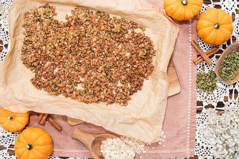A full tray of freshly baked Pumpkin Seed Brittle on parchment paper surrounded by small decorative pumpkins, oats, and cinnamon sticks for a cozy fall aesthetic.