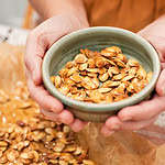 Hands holding a small green bowl filled with golden roasted pumpkin seeds, with a tray of more roasted seeds in the background.