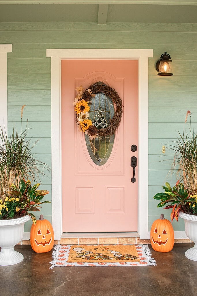 A sage green cottage porch with a pink front door decorated with a grapevine sunflower wreath, flanked by white urn planters filled with ornamental grass and croton, glowing jack-o-lanterns, and a Snoopy Halloween doormat on a pumpkin-print rug.