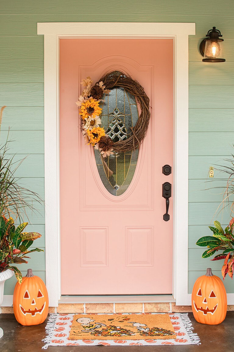 A pink cottage front door decorated with a grapevine sunflower wreath on sage green siding, flanked by white urn planters with ornamental grass and croton, glowing jack-o-lanterns on either side, and a Snoopy Halloween doormat layered on a pumpkin-print rug.