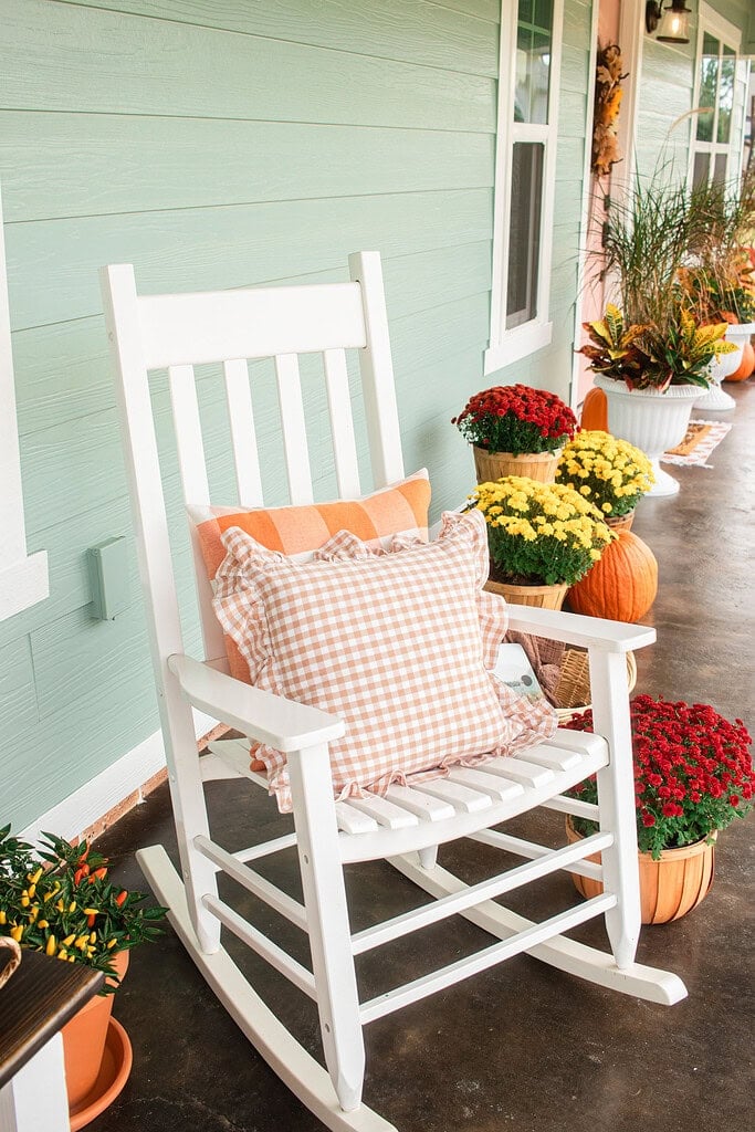 A white rocking chair with a pink gingham ruffle pillow and an orange plaid pillow on a sage green cottage porch, with red and yellow mums in bushel baskets, orange pumpkins, and white urn planters lining the porch behind it.