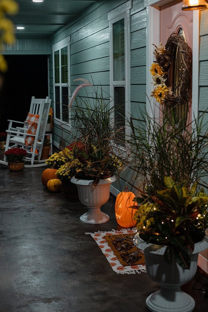 A sage green cottage porch decorated for fall at night, with glowing jack-o-lanterns, white urn planters filled with ornamental grass and croton lit with fairy lights, the pink front door with a sunflower grapevine wreath, and white rocking chairs with fall pillows in the background.