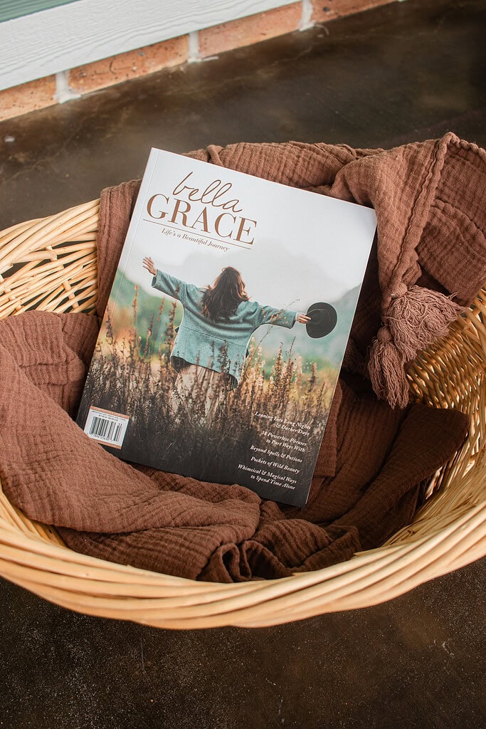 A Bella Grace magazine nestled in a woven basket with a folded brown gauze blanket on a cottage porch, styled as a cozy fall reading nook.