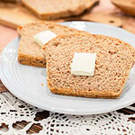 Two slices of fresh milled multigrain seeded sourdough sandwich bread on a light gray plate, each topped with a pat of butter, with the sliced loaf on a wood cutting board blurred in the background and a wooden bowl on a pink linen surface with crochet lace.