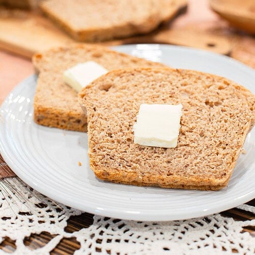 Two slices of fresh milled multigrain seeded sourdough sandwich bread on a light gray plate, each topped with a pat of butter, with the sliced loaf on a wood cutting board blurred in the background and a wooden bowl on a pink linen surface with crochet lace.