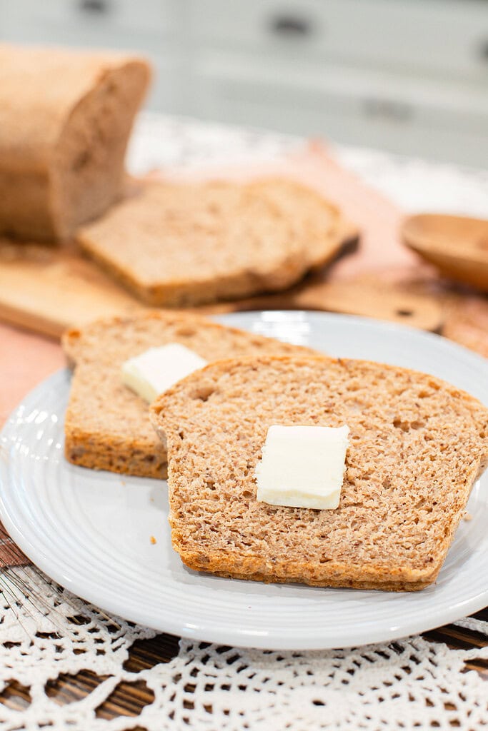 Two slices of fresh milled multigrain seeded sourdough sandwich bread on a light gray plate, each topped with a pat of butter, with the sliced loaf on a wood cutting board blurred in the background and a wooden bowl on a pink linen surface with crochet lace.