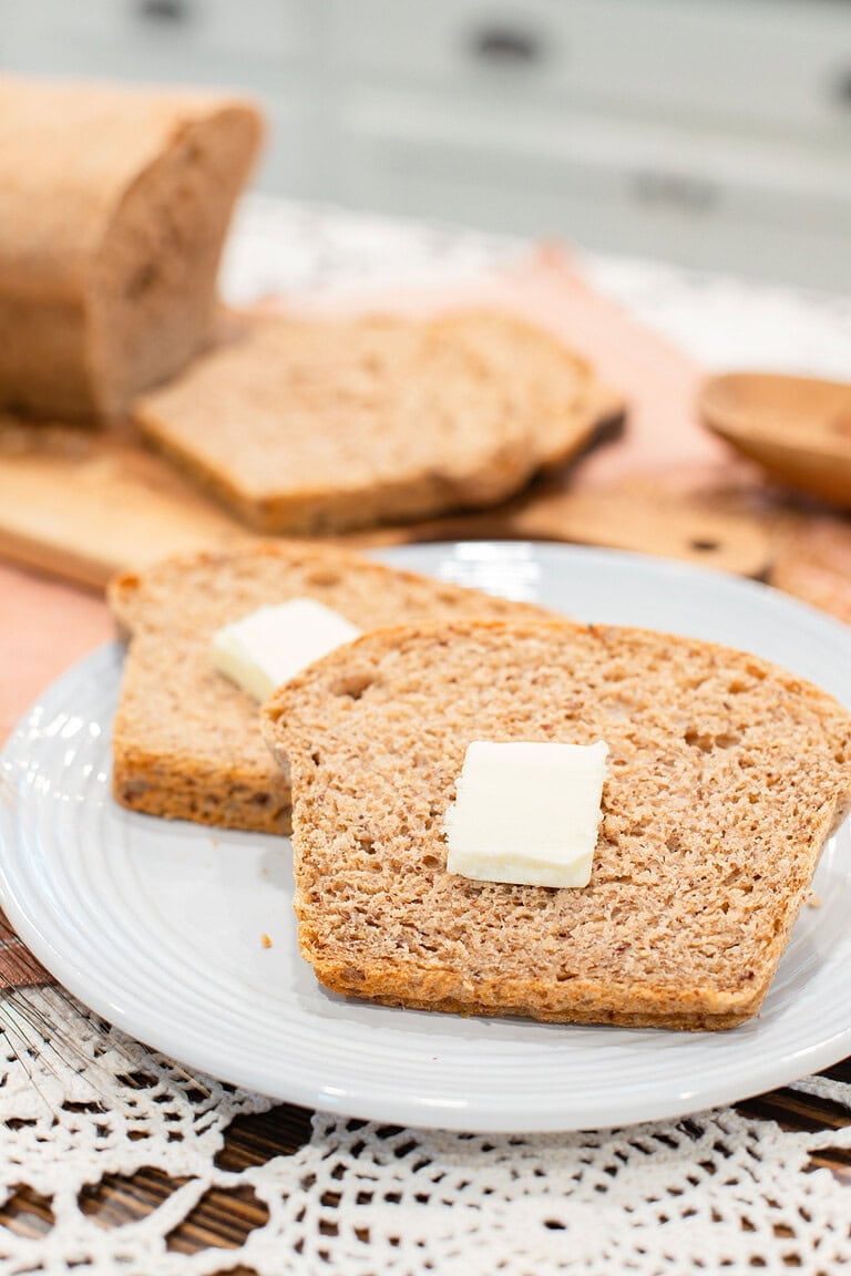 Two slices of fresh milled multigrain seeded sourdough sandwich bread on a light gray plate, each topped with a pat of butter, with the sliced loaf on a wood cutting board blurred in the background and a wooden bowl on a pink linen surface with crochet lace.